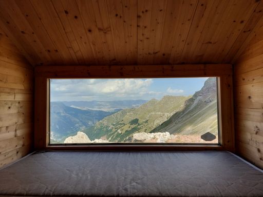 A scenery of the mountains from a panoramic window of a wooden room with a mattress