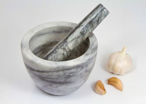 A studio shot of a marble stone mortar with pestle next to whole and peeled garlic on a white table