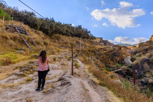 A woman in her 20s walks along a rugged trail through a scenic landscape with blue sky and clouds, showcasing natural beauty and adventure.