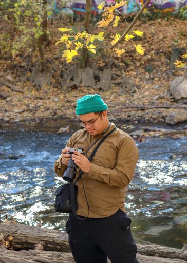 A vertical shot of a guy looking at his vintage camera on a riverbank in an autumn park