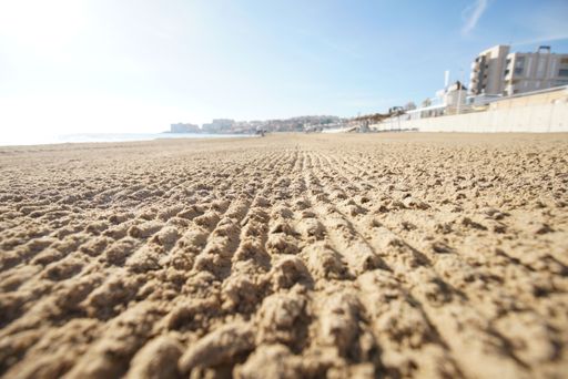 Low angle view of a sandy beach with tire tracks, modern buildings in the background under a clear blue sky, depicting a coastal urban scene.