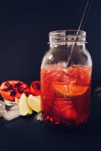 A selective vertical shot of a glass jar cup filled with pomegranate juice with a straw on a black background