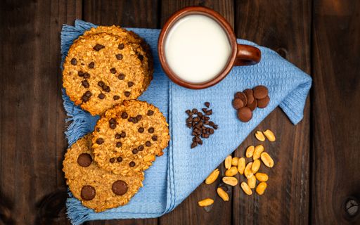 Top view of chocolate chip cookies with a cup of milk on a blue cloth, surrounded by chocolate chips and nuts on a wooden table.