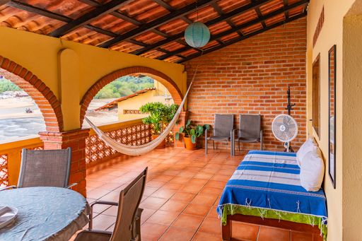 A cozy terrace with hammock, chairs, and a fan overlooking a scenic beach view, featuring brick and tile elements in a tropical setting.