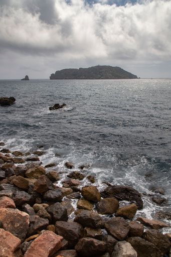 Rocky coastline with waves crashing against the shore under a cloudy sky, distant island visible on the horizon.