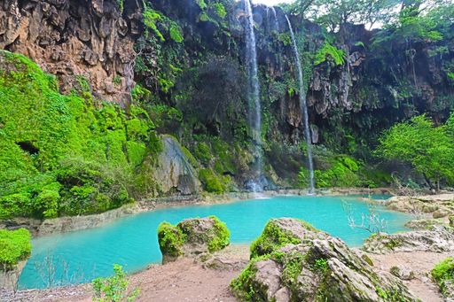 A serene waterfall cascading into a turquoise pool surrounded by lush greenery and rocky cliffs in a natural landscape.