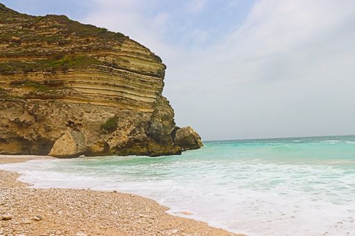 A serene beach scene with turquoise waves gently lapping against a rocky cliff under a partly cloudy sky.