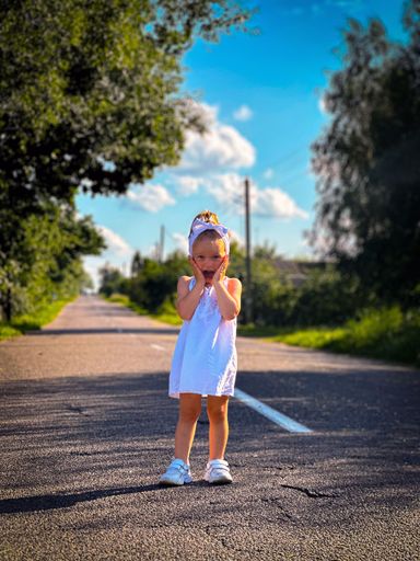 A young girl in a white dress stands on a rural road under a clear blue sky, surrounded by lush greenery.