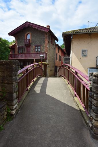 A charming European village scene with a stone bridge leading to rustic houses under a partly cloudy sky.