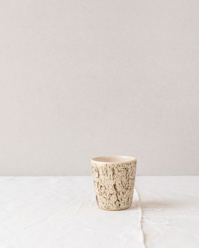 A rustic ceramic cup with a speckled design on a white textured surface, minimalist style.