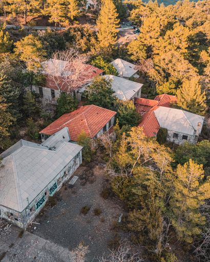 Aerial view of abandoned buildings surrounded by dense forest in a rural area during autumn, showcasing red and grey roofs.