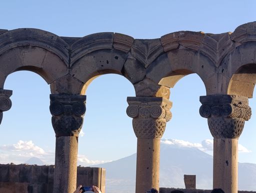 Ancient stone arches with intricate carvings framing a distant mountain landscape under a clear blue sky.