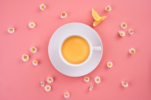 A high angle shot of a cup of orange juice surrounded by small daisy flowers on a pink surface