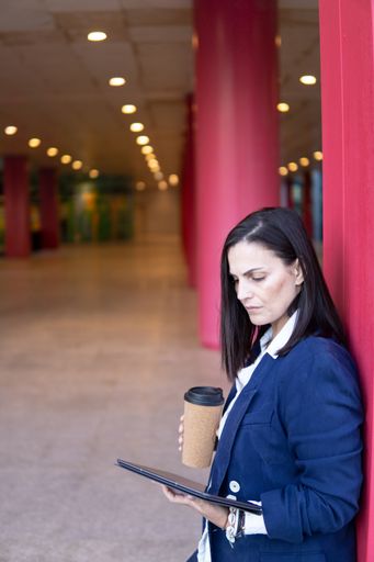 Beautiful Caucasian businesswoman with a cup of coffe and tablet working in the street outside of an office building, vertical shot, copy space