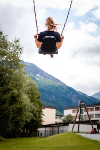 A vertical selective focus shot of a girl from behind swinging in a playground surrounded by mountains