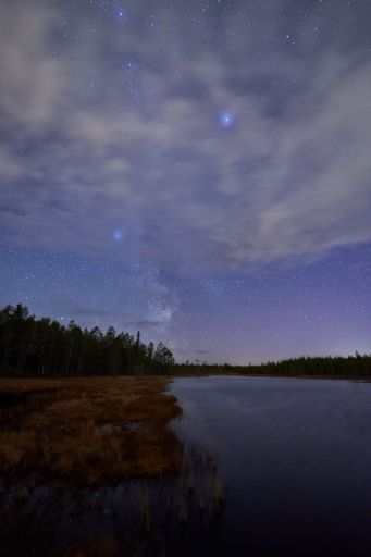 A stunning night sky over a tranquil lake with visible stars and clouds, surrounded by a forest.