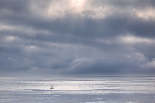 A solitary paddleboarder on a serene ocean under dramatic cloudy skies, creating a tranquil scene.