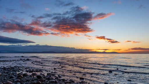 A serene coastal sunset with orange clouds reflecting over a calm sea and pebbled shore.