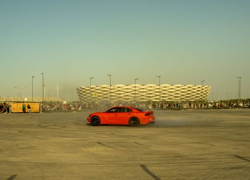 A vibrant red sports car performs a drift in front of a modern stadium with a crowd of spectators under a clear sky.