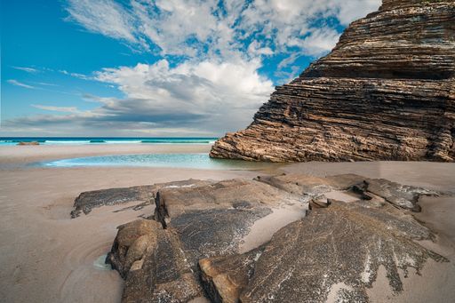 A stunning beach scene with layered rock formations and a clear blue sky, featuring gentle waves and sandy shores.