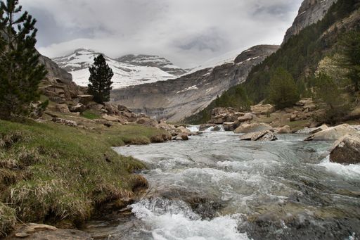 Scenic view of a mountain stream flowing through grassy terrain with snow-capped peaks in the background, under a cloudy sky.