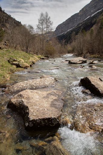 Serene mountain river flowing through a rocky landscape with lush greenery and distant cliffs under a cloudy sky.