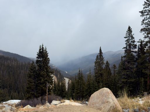 Mountain road through evergreen forest with rocky foreground snow patches and misty distant peaks