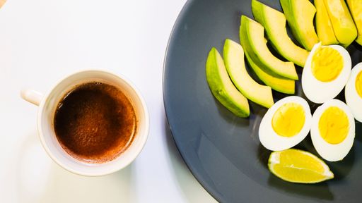 A top view of a breakfast setting with a cup of coffee, sliced avocado, boiled eggs, and a lime wedge on a plate.