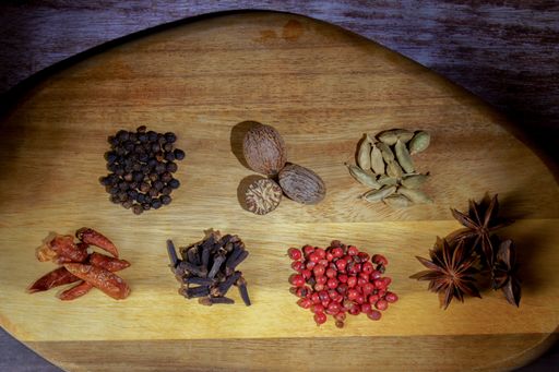 Various whole spices including peppercorns, nutmeg, cardamom, chili, cloves, pink pepper, and star anise on wood