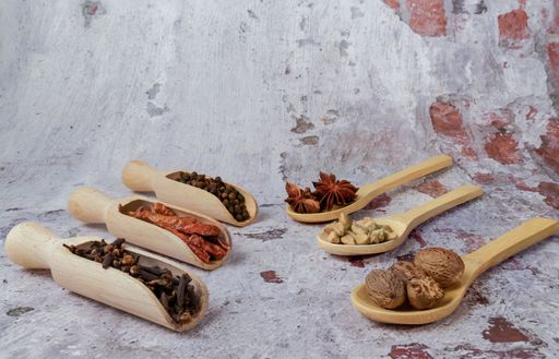 Various spices and herbs including nutmeg, star anise, cardamom, peppercorns, chili peppers, and cloves in wooden spoons