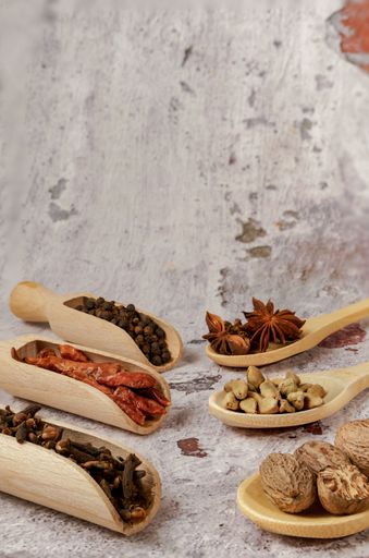 Various spices and herbs including nutmeg, star anise, cardamom, peppercorns, chili peppers, and cloves in wooden spoons