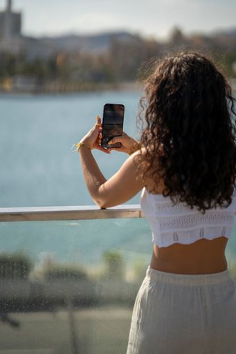 A young woman with curly hair takes a photo with her smartphone by the waterfront on a sunny day.