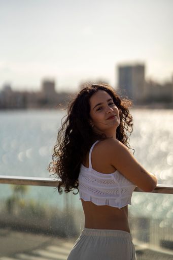 Young woman with curly hair in a white top enjoying a sunny day by the waterfront with a cityscape in the background.