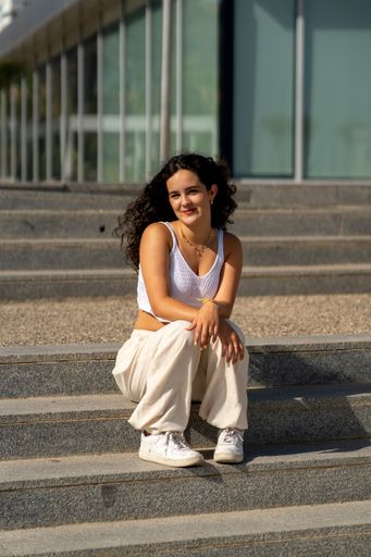Young woman sitting on steps outside a modern building, wearing casual white attire, enjoying a sunny day.