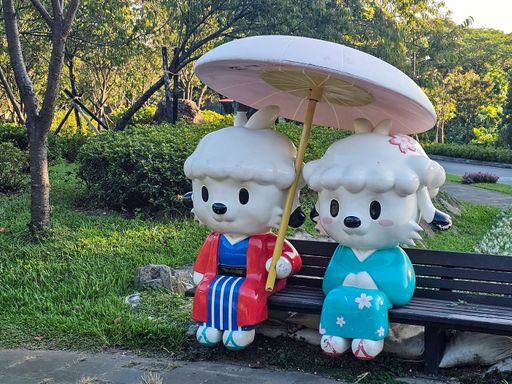 Cute cartoon sheep statues in colorful traditional attire sit on a park bench under a parasol, surrounded by lush greenery.