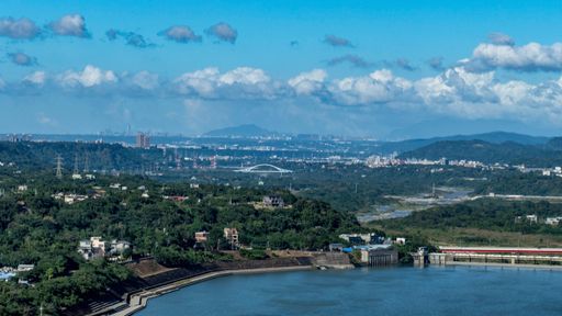 Scenic view of a city skyline with lush green hills and a river under a bright blue sky, featuring a bridge in the distance.