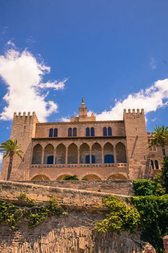 A view of the Royal Palace of La Almudaina in Palma de Mallorca, Spain, under a clear blue sky with fluffy white clouds.