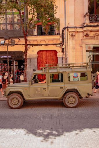 A green military-style vehicle is parked on a sunlit street in front of a European-style building.