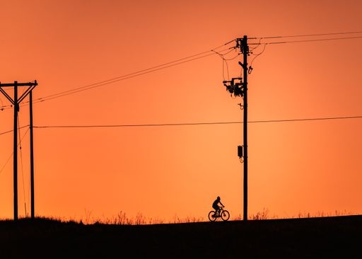 The silhouette of a man on a bike and electric poles against an orange sky at dusk