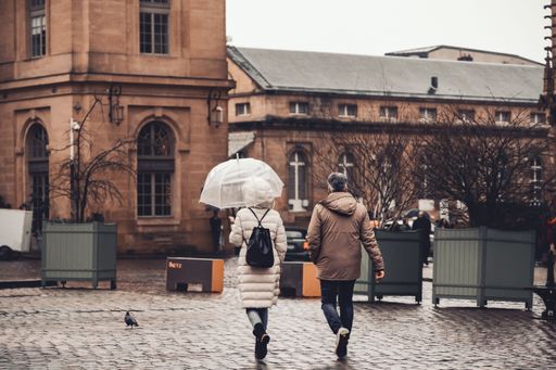 A couple walks across a wet cobblestone square in Metz, with historic architecture visible.
