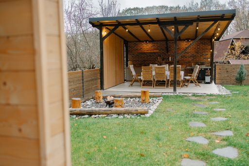Outdoor patio with wooden chairs and table under a modern pergola, surrounded by green grass and stepping stones.