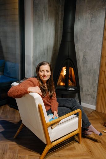 A woman in her 30s relaxes in a cozy chair by a modern fireplace indoors, wearing a warm sweater.