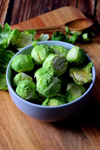 Fresh Brussels sprouts in a bowl on a wooden cutting board with a wooden spoon, showcasing healthy eating and cooking ingredients.