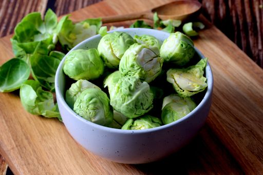 Fresh Brussels sprouts in a white bowl on a wooden surface, with scattered leaves and a wooden spoon in the background.
