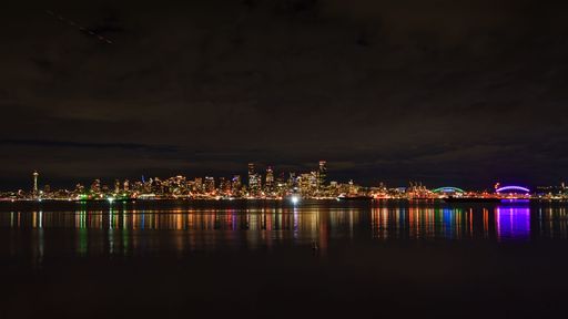 Seattle skyline at night with vibrant city lights reflecting on the calm water under a cloudy sky.