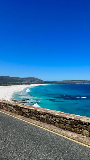 Scenic coastal view of a pristine beach with turquoise waters and a clear blue sky along a stone wall road.