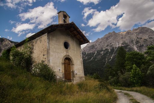 A rustic chapel set against a dramatic mountain backdrop under a partly cloudy sky, capturing the serene beauty of nature.