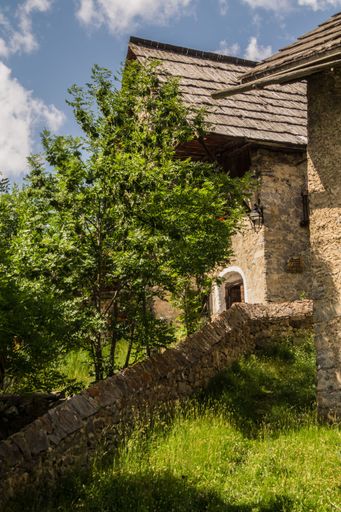 Rustic stone building with lush greenery and clear blue sky in a scenic village setting.