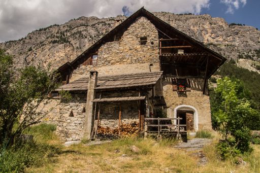 Charming rustic stone house with wooden accents set against a mountainous backdrop under a partly cloudy sky.