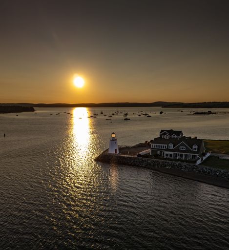 A scenic sunset view over a calm harbor with a lighthouse and houses in the foreground, reflecting golden hues on the water.